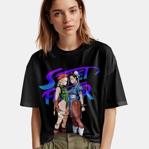 May include: Black t-shirt featuring a graphic of two female characters from the Street Fighter video game, with the game's logo in purple and blue. One character wears a green bodysuit, and the other wears a blue dress.