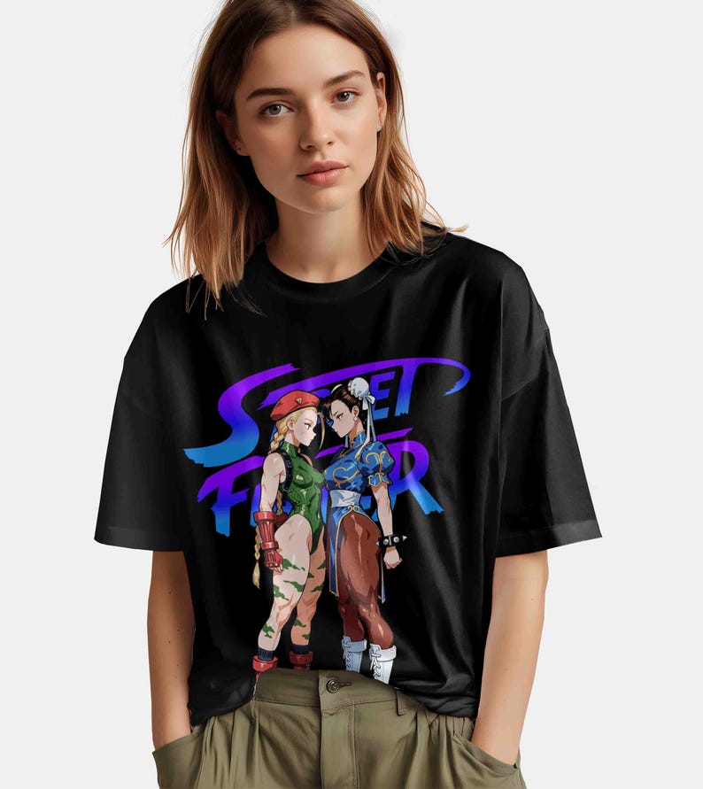 May include: Black t-shirt featuring a graphic of two female characters from the Street Fighter video game, with the game's logo in purple and blue. One character wears a green bodysuit, and the other wears a blue dress.