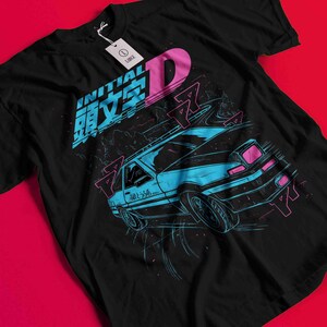 May include: Black t-shirt featuring a blue and pink graphic of a car drifting on a road. The design includes Japanese text and the word "INITIAL D" in pink and blue. A white tag is attached to the shirt.
