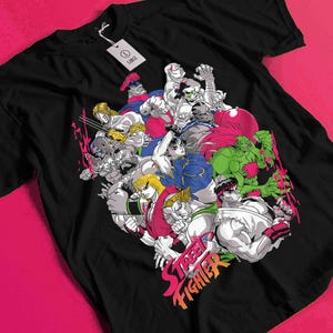 Puede incluir: Camiseta negra con un gráfico colorido de personajes de Street Fighter. El diseño presenta múltiples personajes en poses de acción, con las palabras "Street Fighter" en una fuente estilizada. Una etiqueta blanca está adjunta.