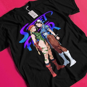 May include: Black t-shirt featuring a graphic of two female characters from the Street Fighter video game series. The design includes the word "Street Fighter" in a purple and blue gradient. The characters are in fighting poses.