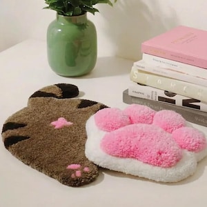 May include: A brown cat-shaped rug with black stripes and a pink star detail, next to a white and pink paw-shaped rug. A green vase with greenery and a stack of books are also in the image.