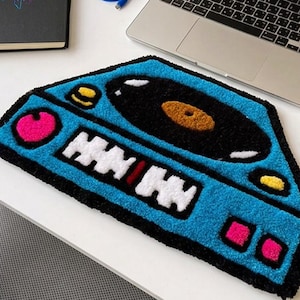 May include: A tufted rug shaped like a DJ turntable console in bright blue, featuring a black vinyl record with an orange center, white fader controls, and pink and yellow accent buttons, all outlined in a thick black border.