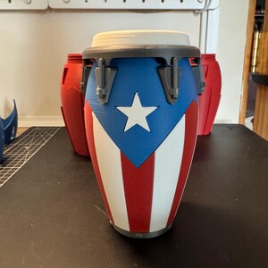 May include: A conga drum featuring a design inspired by the Puerto Rican flag with a blue triangle, a white star, and alternating red and white vertical stripes. The drum has a grey rim with tension hardware and a white drumhead on top.
