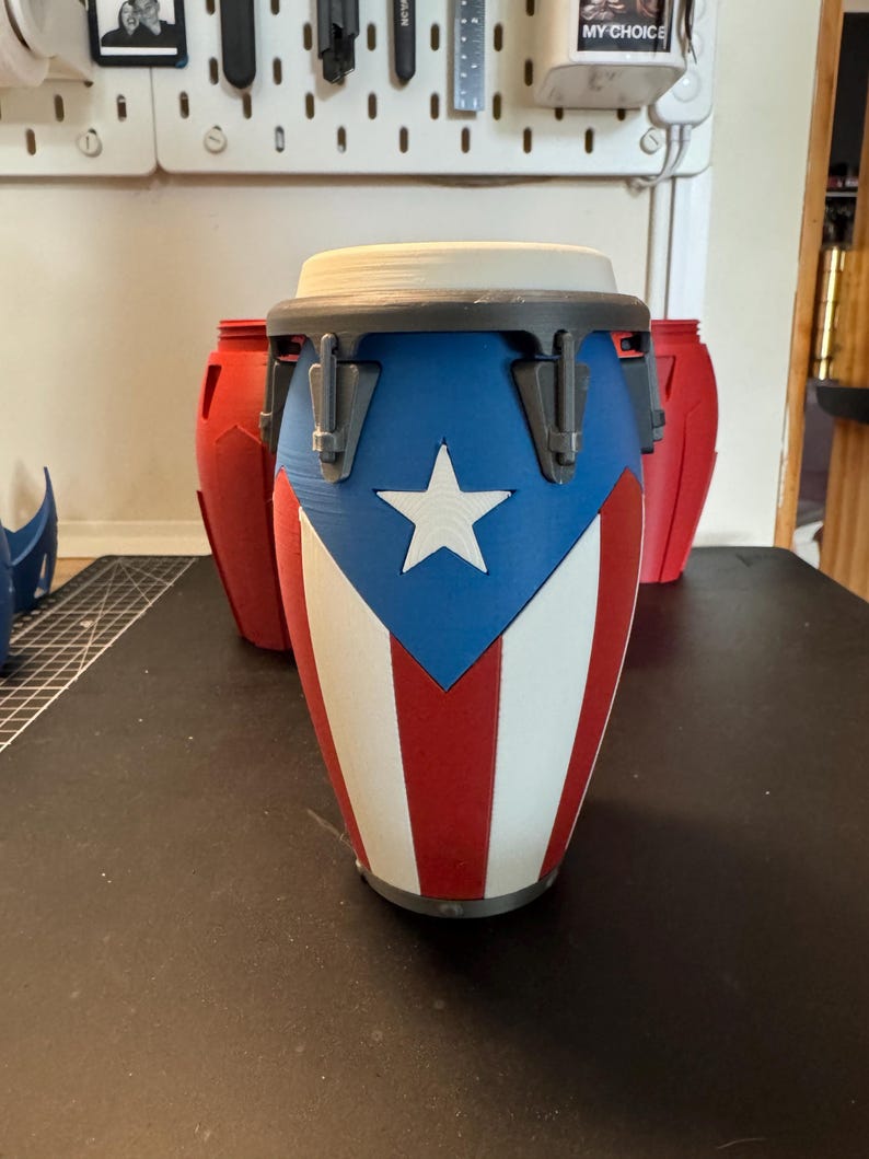 May include: A conga drum featuring a design inspired by the Puerto Rican flag with a blue triangle, a white star, and alternating red and white vertical stripes. The drum has a grey rim with tension hardware and a white drumhead on top.