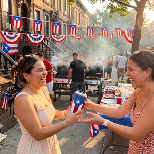 May include: A cylindrical beverage cooler featuring the Puerto Rican flag design with a white star on a blue triangle and red and white stripes. The cooler is held between two individuals outdoors during a street celebration with flags.