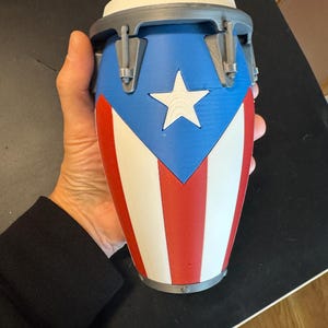 May include: A small conga drum featuring the Puerto Rican flag design with a blue triangle, a white star, and alternating red and white vertical stripes. The drum has a grey rim and hardware, with a white drumhead on top, constructed from plastic.
