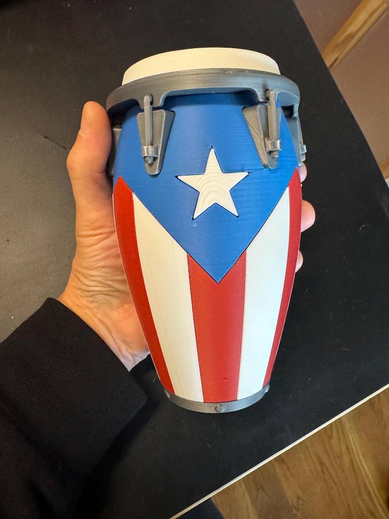 May include: A small conga drum featuring the Puerto Rican flag design with a blue triangle, a white star, and alternating red and white vertical stripes. The drum has a grey rim and hardware, with a white drumhead on top, constructed from plastic.