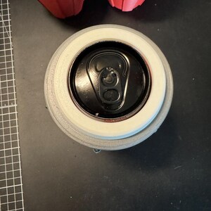 May include: A top-down view of a black aluminium beverage can inserted into a light grey, circular 3D-printed holder. The can features a black pull tab and a visible rim. Two red, star-shaped 3D-printed objects are partially visible at the top.