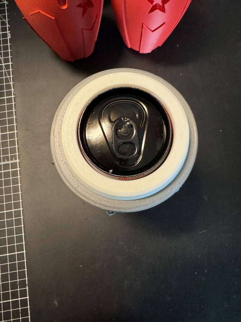 May include: A top-down view of a black aluminium beverage can inserted into a light grey, circular 3D-printed holder. The can features a black pull tab and a visible rim. Two red, star-shaped 3D-printed objects are partially visible at the top.