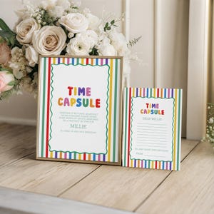 Op de afbeelding: Een ingelijst "Time Capsule"-bord en een bijpassende kaart. Het bord en de kaart hebben een kleurrijke gestreepte rand en de woorden "Time Capsule" in regenboogkleuren. Het bord bevat tekst voor een verjaardagsbericht.
