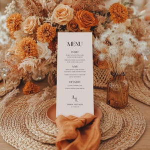 May include: A wedding menu card with the word "MENU" at the top, featuring a list of courses. The card is placed on a woven placemat with a terracotta plate and orange napkin. The background includes a floral arrangement with orange and cream-colored flowers.