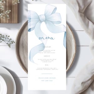 May include: A baby shower menu card featuring a light blue watercolour bow illustration. The menu lists savoury, sweet, and tea options, with the event details: Olivia's Baby Shower, May 23rd 2026.
