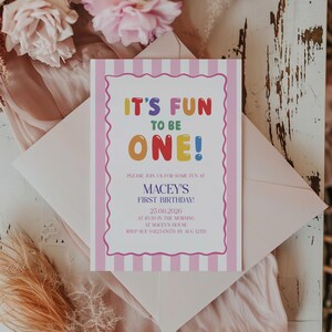 Op de afbeelding: Een rechthoekige verjaardagsuitnodiging met een roze en wit verticaal gestreepte rand en een geschulpt wit middenstuk. De tekst luidt: IT'S FUN TO BE ONE! PLEASE JOIN US FOR SOME FUN AT MACEY'S FIRST BIRTHDAY! 25.08.2026 AT 10:30 IN THE MORNING AT MACEY'S HOUSE RSVP SUE 0412345678 BY AUG 12TH.