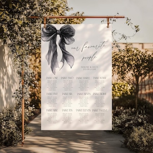 May include: A white wedding seating chart with a watercolor-style black bow illustration. The text reads "our favourite people" and lists names under table numbers. The wedding date is displayed as 25.08.2020. The sign is suspended from a copper-colored rod.