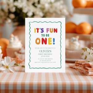 Op de afbeelding: Een witte uitnodigingskaart met een groene geschulpte rand. De tekst "IT'S FUN TO BE ONE!" is geschreven in kleurrijke letters. De uitnodiging luidt "OLIVER'S FIRST BIRTHDAY! 25.08.2026".