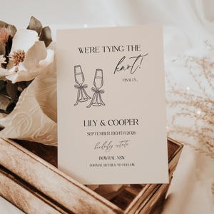 May include: A cream-colored wedding invitation with black script. The text reads "WE'RE TYING THE knot!" with the names "LILY & COOPER" and the date "SEPTEMBER EIGHTH 2026". Two champagne glasses are illustrated.
