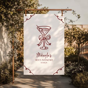 May include: A white banner featuring a maroon cocktail illustration and text. The banner reads "Mikayla's HEN'S WEEKEND! 07.08.26" in a decorative font. The banner is suspended from a copper-coloured rod.