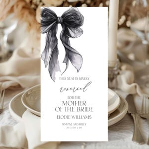 Op de afbeelding: Een wit naambordje met een zwarte strik in aquarel-stijl bovenaan. De tekst luidt "THIS SEAT IS KINDLY reserved FOR THE MOTHER OF THE BRIDE" met de namen Elodie Williams, Simone en Riley en de datum 25 | 06 | 20.