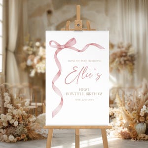 May include: A white sign with a pink ribbon bow and the text "Thank you for celebrating Ellie's First Bowtiful Birthday April 22nd 20XX".