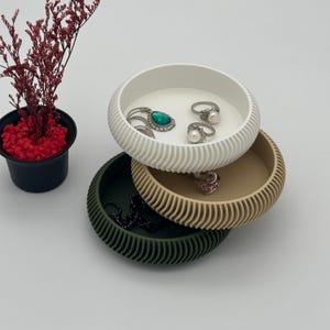 Könnte beinhalten: Drei übereinander gestapelte runde Schmuckschalen mit einem strukturierten Design. Die oberste Schale ist weiß und enthält einen türkisfarbenen Ring, einen silbernen Ring mit einer Perle und ein Paar silberne Ohrringe. Die mittlere Schale ist beige und die unterste Schale ist grün.