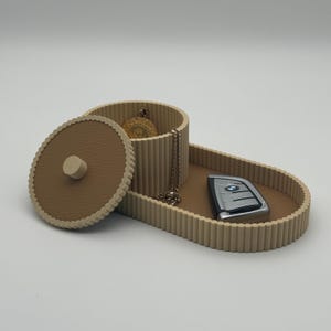 Può includere: Un organizzatore beige e marrone con un coperchio e un vano separato. L'organizzatore ha una texture a coste e  è progettato per contenere piccoli oggetti come gioielli e chiavi.