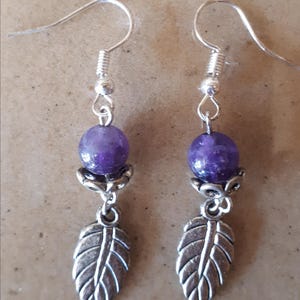 Puede incluir: Un par de pendientes colgantes con una cuenta redonda de color púrpura oscuro, un adorno floral plateado y un dije de hoja plateada. Los pendientes tienen ganchos para las orejas plateados.