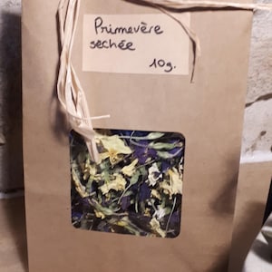 Op de afbeelding: Een bruine papieren zak, vastgebonden met raffia, met gedroogde sleutelbloem bloemen. Een rechthoekig venster onthult de gele en paarse bloemen. Het etiket luidt "Primavère séchée 10g."