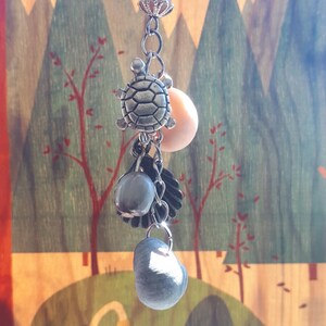 Peut inclure: Un charm argenté avec une tortue, des perles et d'autres éléments décoratifs. Le charm est suspendu à un fermoir. Le fond présente un panneau en bois avec une scène de paysage peinte.