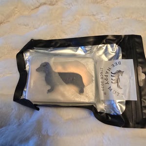 Könnte beinhalten: Eine transparente Plastiktüte mit einem schwarzen Reißverschluss und einem weißen Etikett, auf dem "Happy Mutt" und "Beef" mit einer Hundesilhouette steht. In der Tüte befindet sich eine weiße Seifenstange mit einer schwarzen Silhouette eines Dackels.