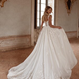 Op de afbeelding: Een witte bruidsjurk met een strakke buste en een volle rok. De jurk is versierd met delicate bloemenborduursels en een lange sleep.