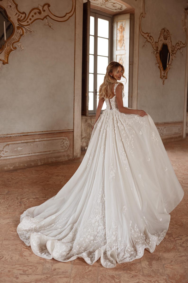 Op de afbeelding: Een witte bruidsjurk met een strakke buste en een volle rok. De jurk is versierd met delicate bloemenborduursels en een lange sleep.