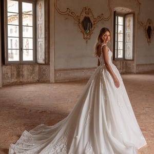 Op de afbeelding: Een bruid in een witte bruidsjurk met een volle rok en delicate bloemenborduursels. De jurk heeft een getailleerd lijfje en een lage rug. De bruid staat in een grote, sierlijke kamer met hoge plafonds en ramen.