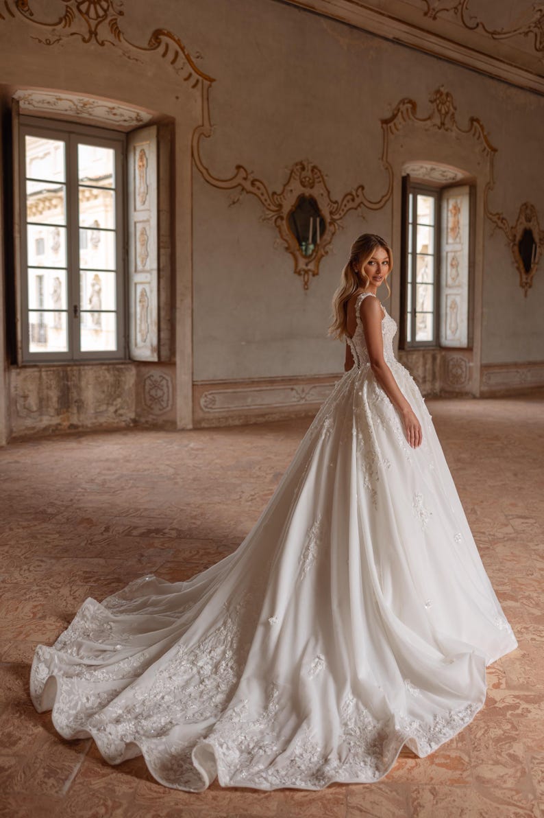 Op de afbeelding: Een bruid in een witte bruidsjurk met een volle rok en delicate bloemenborduursels. De jurk heeft een getailleerd lijfje en een lage rug. De bruid staat in een grote, sierlijke kamer met hoge plafonds en ramen.