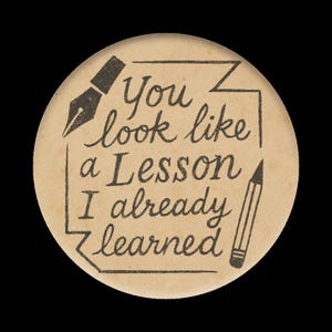 May include: A round, tan button with black text and illustrations. The text reads "You look like a Lesson I already learned." A pen and pencil are drawn on either side of the text.