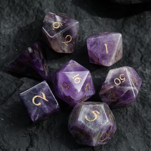 May include: A collection of seven amethyst gemstone dice with gold-colored numbers. The dice are various shapes, including a d4, d6, d8, d10, d12, and d20. The dice are arranged on a dark, textured surface.