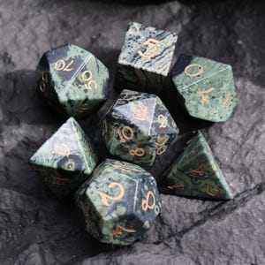 May include: A set of seven polyhedral dice in shades of green and black with gold-colored numbering. The dice are arranged on a dark, textured surface, showcasing their various geometric shapes, including a d4, d6, d8, d10, d12, d20, and a percentile die.