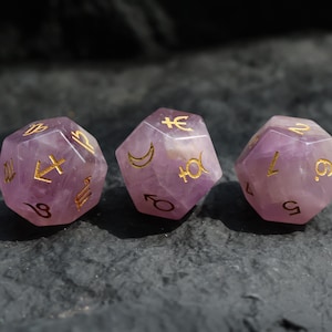 Może przedstawiać: Trzy ametystowe kostki do gry ze złotymi symbolami astrologicznymi i liczbami. Kostki są w jasnofioletowym kolorze i mają fasetowany wzór. Kostki ułożone są w rzędzie na ciemnoszarej powierzchni.