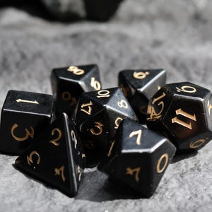 May include: A collection of black polyhedral dice with gold numbering, including D6, D4, D8, D10, D12, and D20. The dice are arranged on a textured gray surface, with some dice partially obscured.