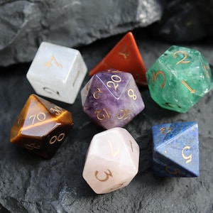 May include: A collection of gemstone dice in various shapes and colors, including a clear quartz d6, a tiger's eye d8, a purple amethyst d20, a green d12, a pink rose quartz d10, a blue lapis lazuli d6, and an orange d4. The dice are set against a dark, textured background.