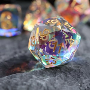 Puede incluir: Un dado d20 iridiscente y multifacético con numeración dorada, apoyado sobre una superficie oscura y texturizada. Otros dados son visibles en el fondo, creando una colección de accesorios de juego. El dado refleja un espectro de colores.