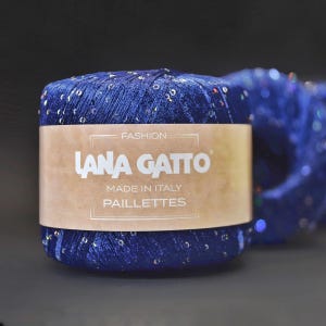Puede incluir: Una madeja de hilo azul con lentejuelas plateadas, etiquetada "Lana Gatto" y "Paillettes" con las palabras "Fashion" y "Made in Italy".