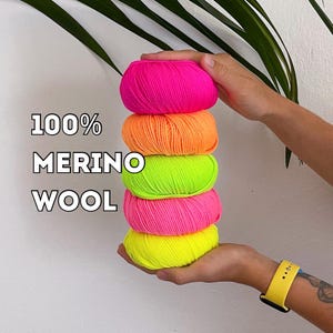 Op de afbeelding: Een stapel van vijf felgekleurde bollen merinowol in neonroze, oranje, groen, roze en geel. De tekst "100% MERINO WOOL" staat over de afbeelding. De wol wordt in de handen van een persoon gehouden.