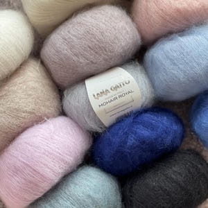 May include: A collection of yarn skeins in various colours, including pink, blue, white, and black. The yarn is soft and fluffy, and the skeins are arranged in a circle. The label on one of the skeins reads "Lana Gatto Mohair Royal Made in Italy www.lanagatto.it".