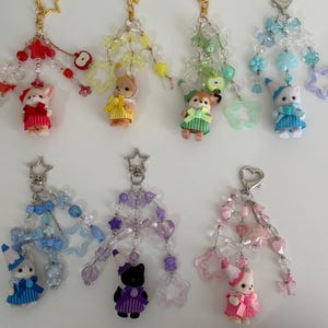 May include: Seven decorative keychains featuring small animal figurines wearing party hats and dresses. Each keychain includes colorful beads, star-shaped charms, and metal clasps in red, yellow, green, blue, purple, and pink color themes.