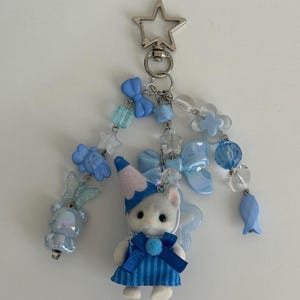 May include: A keychain featuring a small flocked cat figurine wearing a blue striped dress and a blue party hat. The charm is decorated with various light blue and clear beads, including shapes of bows, stars, a butterfly, a bear, and a fish, all attached to a silver-toned star-shaped lobster clasp.