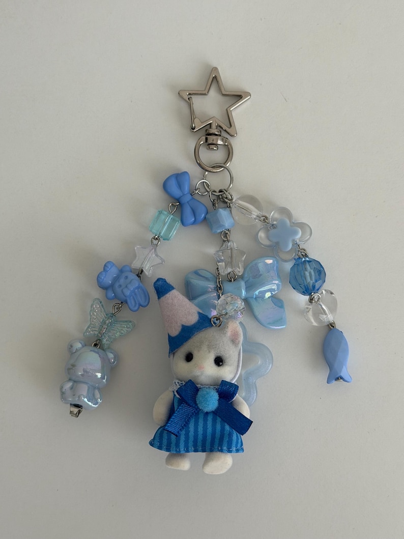 May include: A keychain featuring a small flocked cat figurine wearing a blue striped dress and a blue party hat. The charm is decorated with various light blue and clear beads, including shapes of bows, stars, a butterfly, a bear, and a fish, all attached to a silver-toned star-shaped lobster clasp.