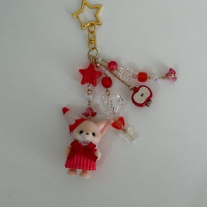 May include: A gold-toned star-shaped keychain featuring a flocked animal figurine wearing a red and white striped dress and hat. The charm includes dangling red and clear beads, a red apple charm, a clear heart, and a small butterfly accent.