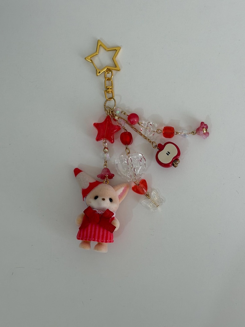May include: A gold-toned star-shaped keychain featuring a flocked animal figurine wearing a red and white striped dress and hat. The charm includes dangling red and clear beads, a red apple charm, a clear heart, and a small butterfly accent.