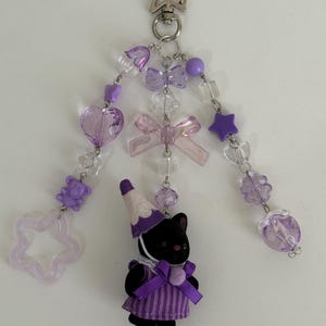 May include: A keychain featuring a star-shaped metal clasp and three dangling strands of purple and clear beads, including heart, star, and bear shapes, plus bows. A small black animal figure wearing a purple striped dress and party hat hangs below.